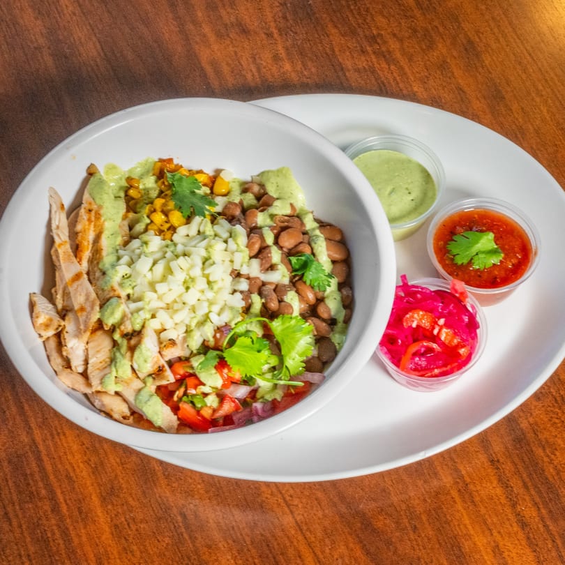 Grilled chicken bowl with black beans, corn, salsa, and green sauce on a wooden table.