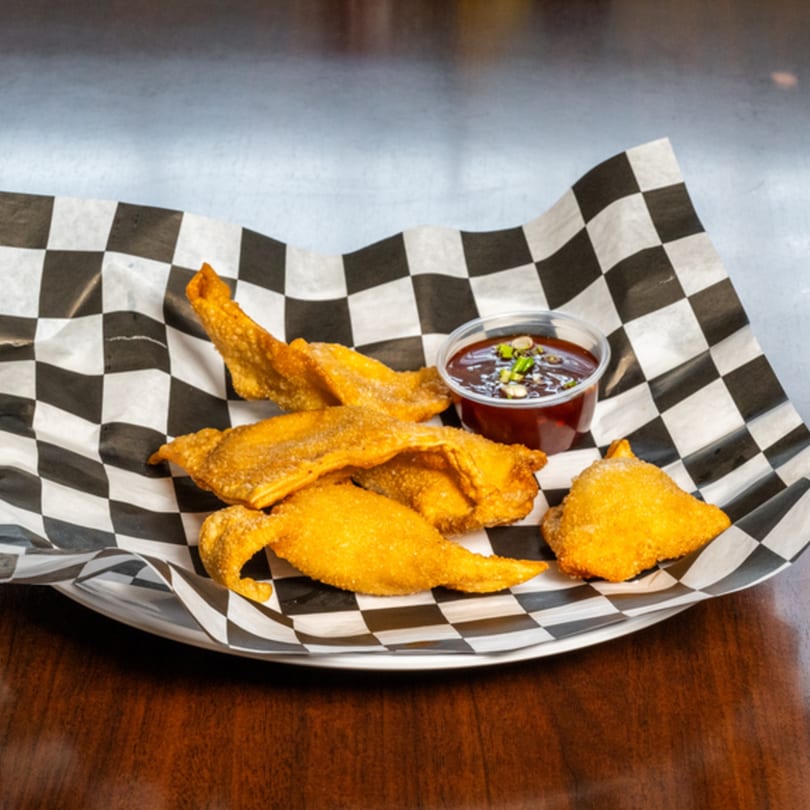 Golden fried wontons and a small cup of dark dipping sauce with green onions on checkered paper.