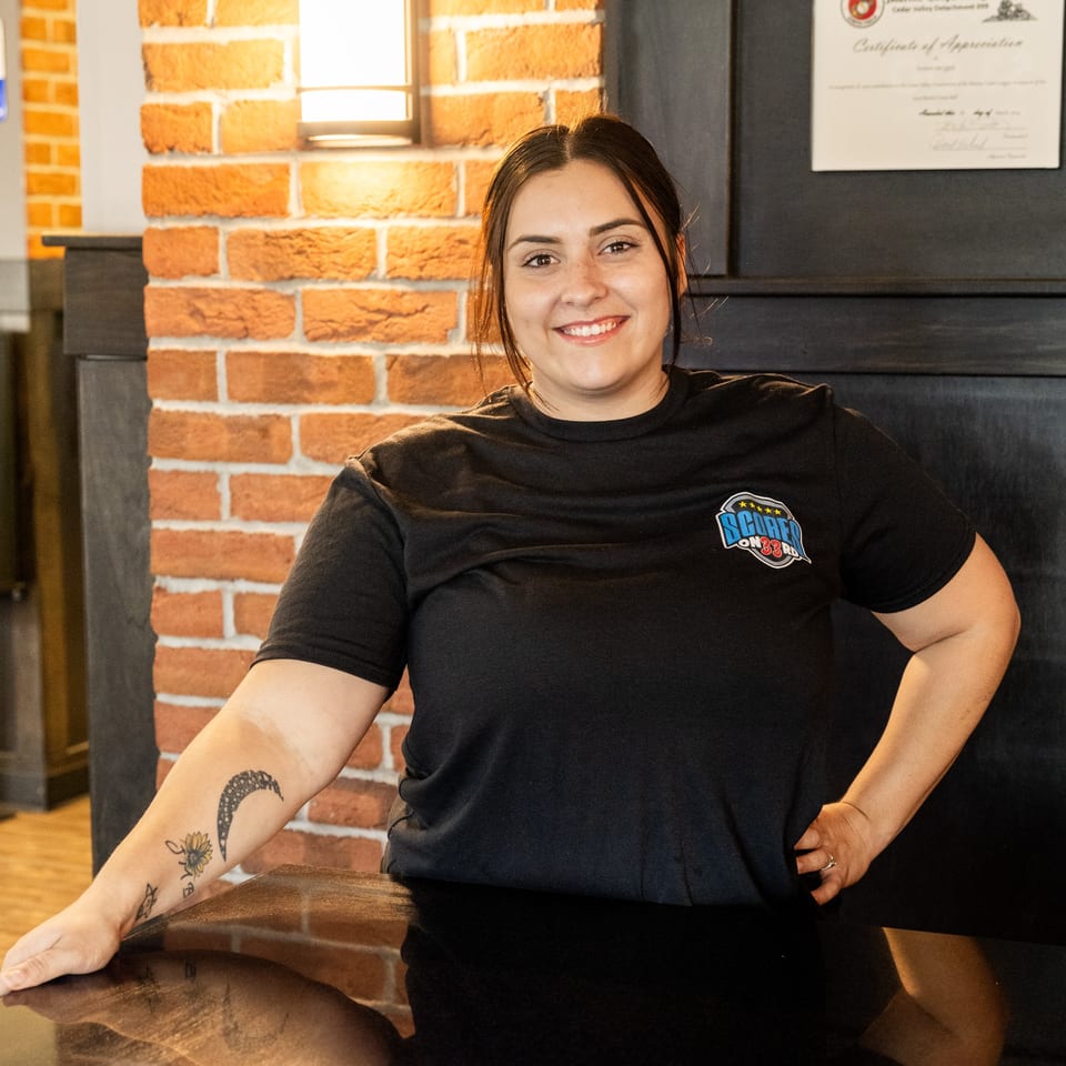 Smiling Scores on 33rd staff member with tattooed arm leaning on a dark counter.