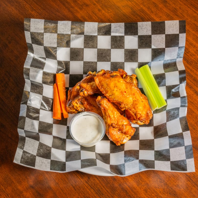 Spicy chicken wings with carrots, celery, and ranch dressing on checkered paper.