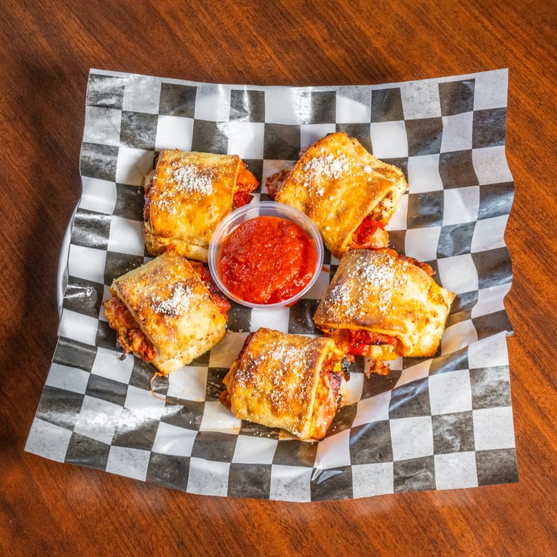 Five golden-brown pizza rolls with powdered sugar and a side of marinara sauce on checkered paper.