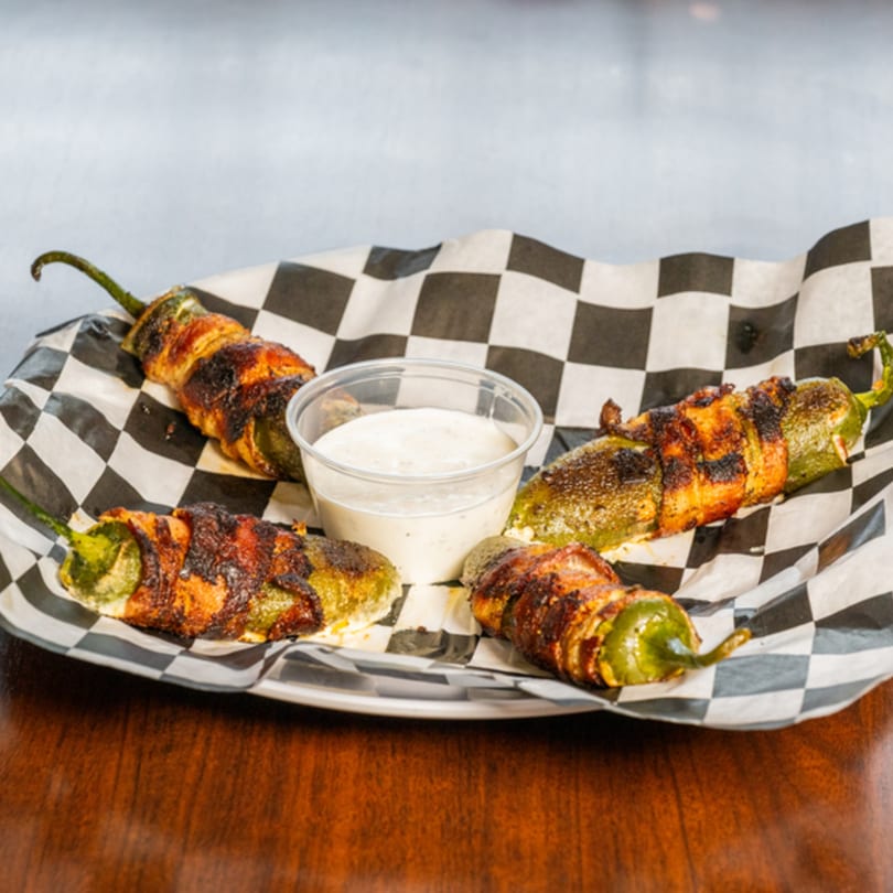 Four bacon-wrapped jalapeño poppers with a side of ranch dressing on checkered paper.