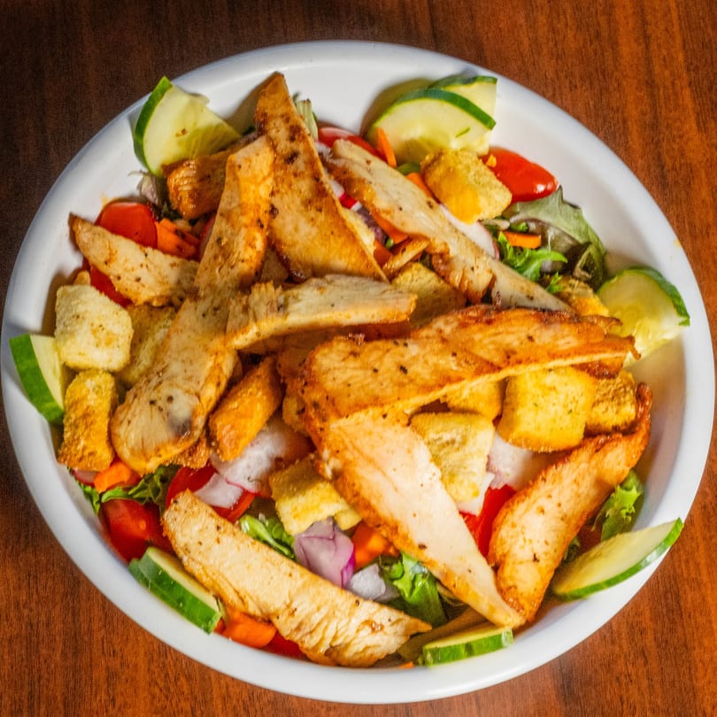 Grilled chicken salad with cucumbers, tomatoes, red onions, and croutons in a white bowl.